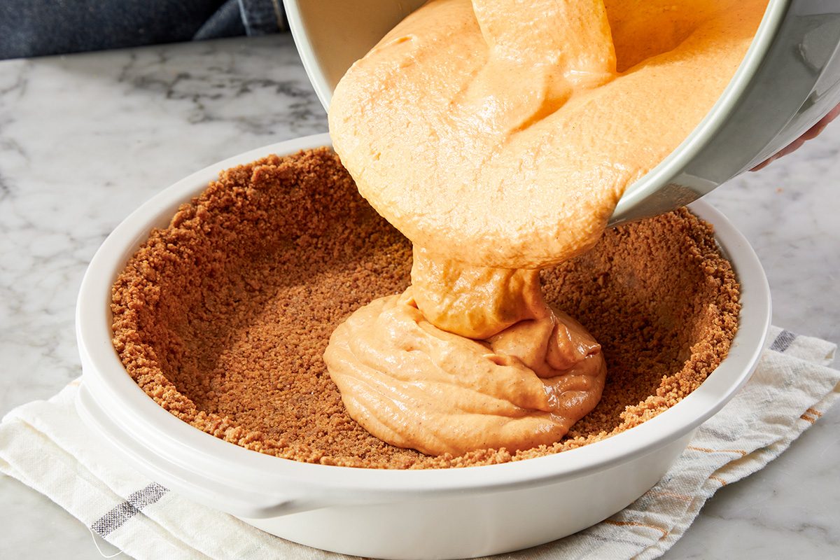 A bowl of orange pumpkin pie filling is being poured into a pie crust in a white dish, which sits on a striped cloth on a marble countertop.