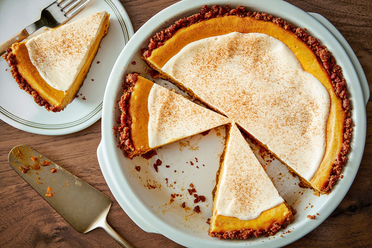 A partially sliced pumpkin pie with a creamy topping and sprinkled cinnamon, displayed in a white pie dish. One slice is served on a plate with a fork beside it. A pie server rests nearby on a wooden table.