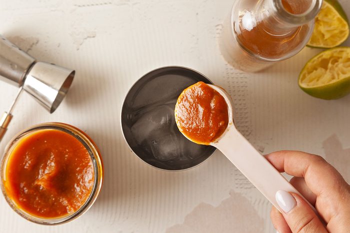 A hand holds a white spoon with a dollop of red sauce above a glass with ice. Nearby are a jar of sauce, a metal jigger, a bottle, and squeezed lime halves on a light surface.