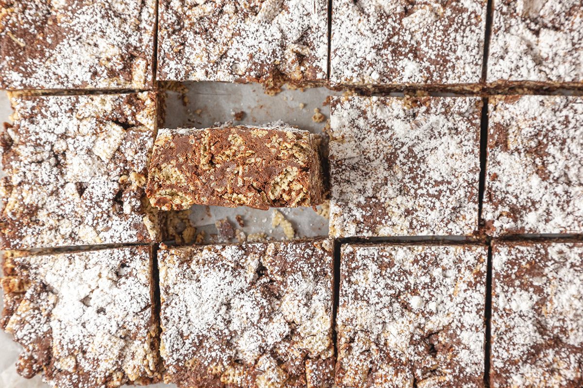 Overhead shot of chocolate oat-based Puppy Chow Bars cut into neat squares, dusted with powdered sugar; One bar is turned on its side to reveal the crunchy texture inside