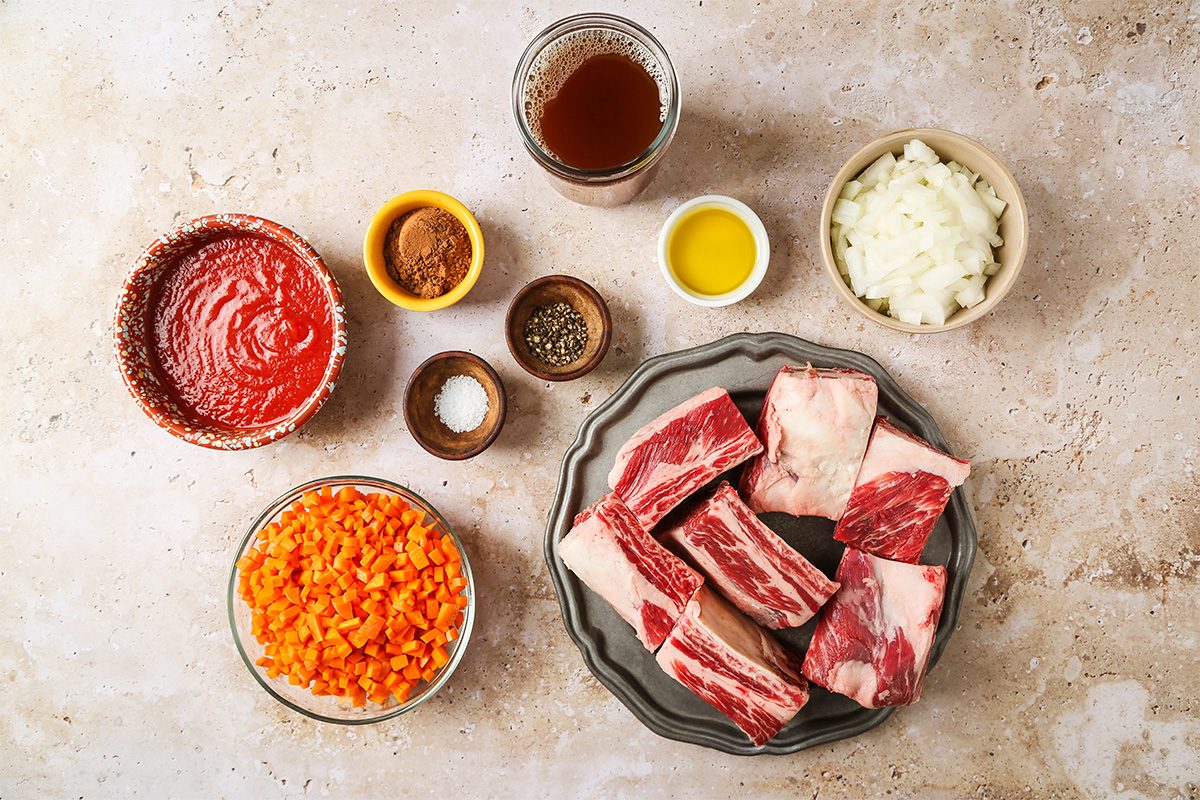 A top-down view of ingredients for a beef dish, including raw beef short ribs, chopped carrots, diced onions, tomato sauce, olive oil, broth, and small bowls of spices arranged on a light surface.