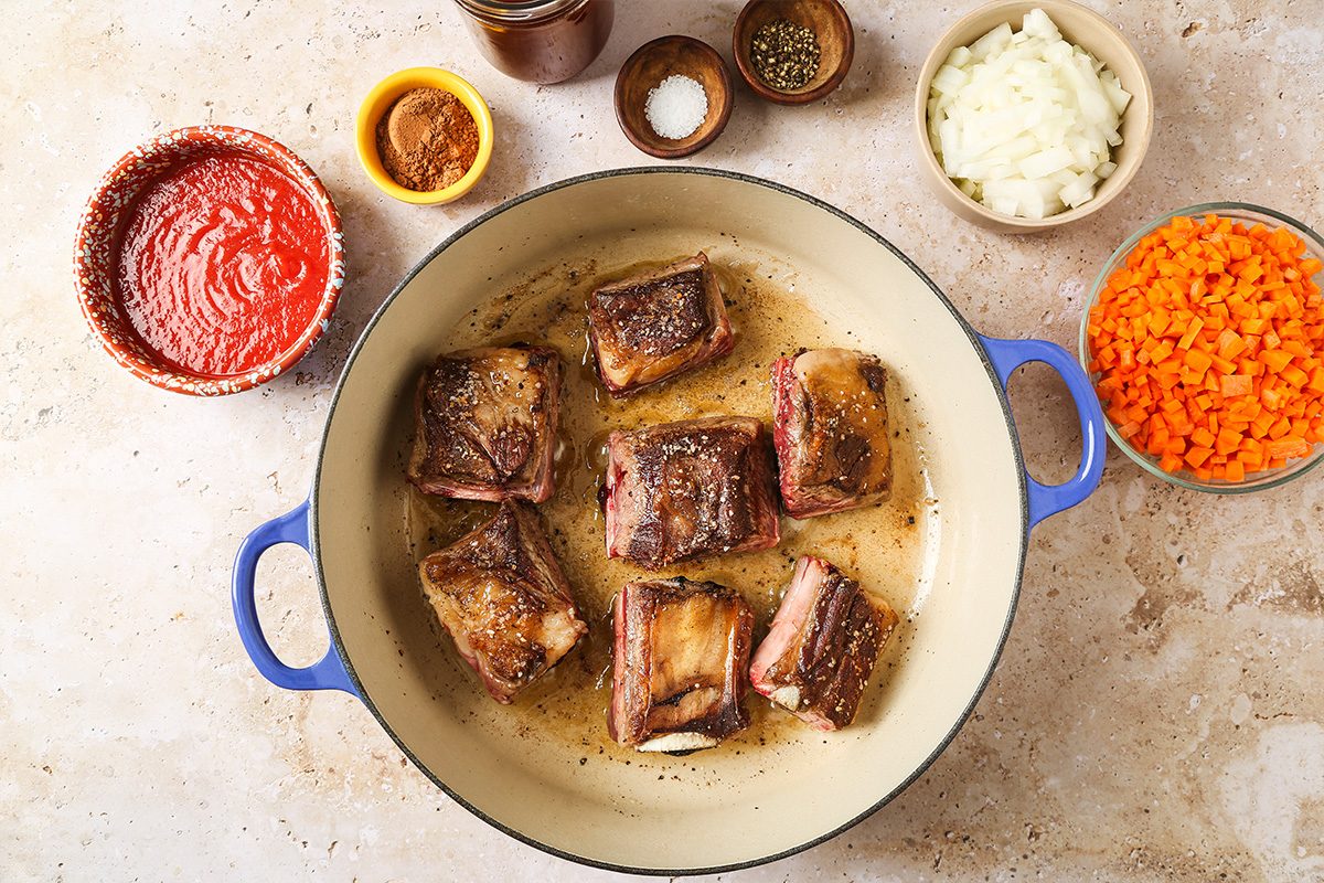 A blue-handled pot with browned beef short ribs, surrounded by bowls of tomato sauce, chopped carrots, onions, spices, and seasonings on a light countertop.