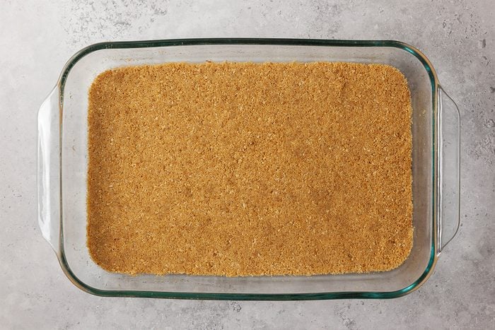 A glass baking dish filled with an even layer of graham cracker crust, pressed down and ready for baking, sits on a light gray countertop.