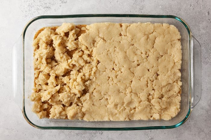 A glass baking dish filled with a crumbly dough mixture, with half of the dough loosely crumbled and the other half pressed flat and smooth, on a light gray surface.
