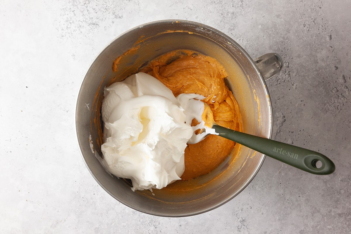 A metal mixing bowl contains pumpkin-colored batter and a large dollop of fluffy whipped egg whites, with a green spatula resting inside. The bowl sits on a light gray countertop.
