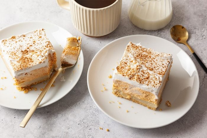 Two plates each with a square slice of layered cake topped with whipped cream and cinnamon, one with a fork cutting a bite. A cup and a small jug, possibly with milk, are in the background.