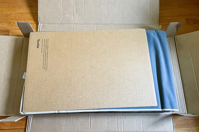 An opened cardboard box with a flat, rectangular piece of cardboard on top and a folded blue fabric partially visible inside. The box is resting on a wooden floor.