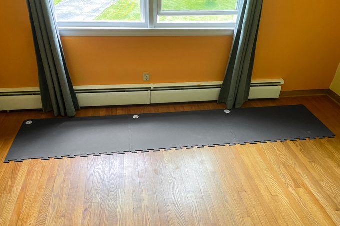 A long black interlocking foam mat lies on a hardwood floor in front of a window with gray curtains. The walls are orange, and there’s a white baseboard heater below the window.