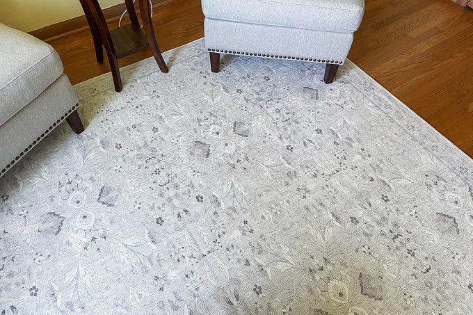 A light gray patterned area rug on a wooden floor, bordered by two light-colored upholstered chairs with dark wooden legs. The rug features subtle floral and geometric designs.