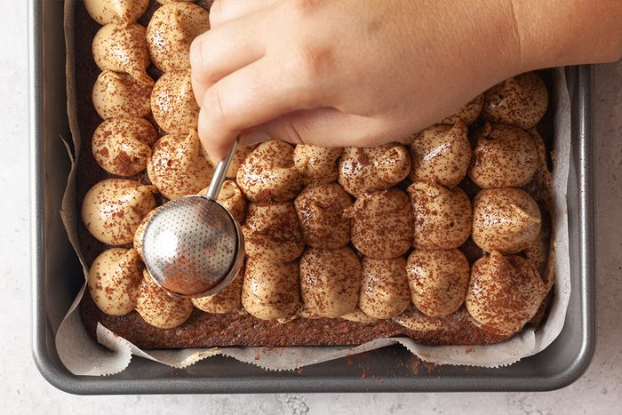 A hand uses a small mesh sifter to dust cocoa powder over piped dollops of cream on a dessert in a rectangular baking pan lined with parchment paper.