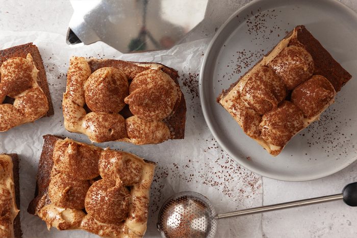 Overhead view of several slices of tiramisu topped with cocoa powder on a white surface, with one slice on a round plate, a metal strainer, and a coffee pot nearby.