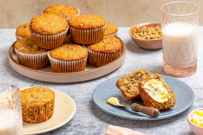 Table view shot of vegan banana muffins; A blue plate holds a halved muffin spread with butter and a knife, with a glass of milk and a bowl of chopped nuts nearby; More muffins in paper liners are stacked on a round tray, with an additional muffin on a beige plate; A small bowl of butter and a peach napkin rest on the gray surface