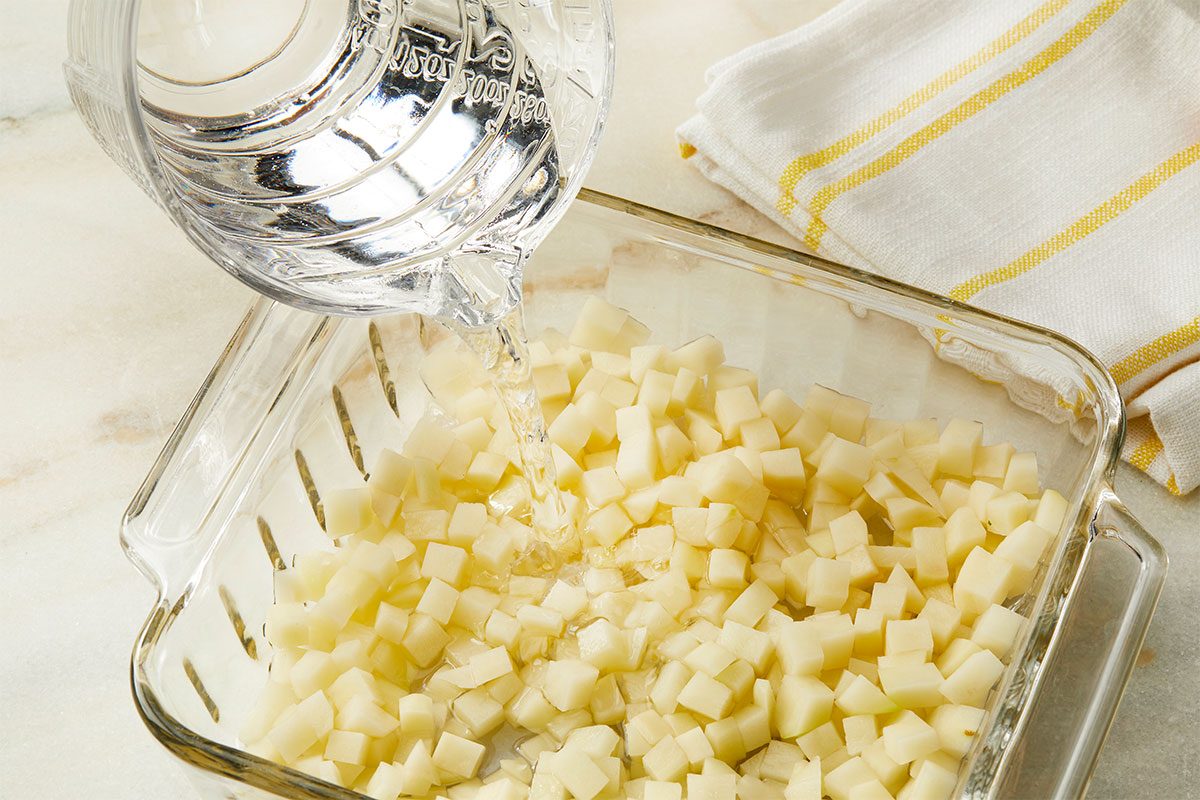 A glass measuring cup pours water over diced potatoes in a clear glass baking dish, with a yellow-striped kitchen towel nearby on a light countertop.