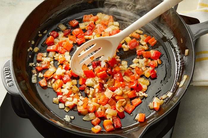 Diced red bell peppers and onions are being sautéed in a black skillet with a white slotted spoon, with steam rising from the vegetables.