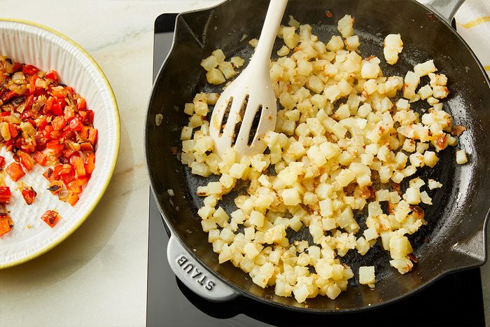 Diced potatoes cooking in a black skillet with a white slotted spoon, next to a bowl of diced cooked vegetables on a counter.