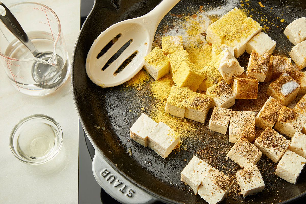 A skillet contains cubed tofu sprinkled with various seasonings. Beside the skillet, a measuring spoon and two small bowls, one with liquid and one empty, are placed on a white surface.