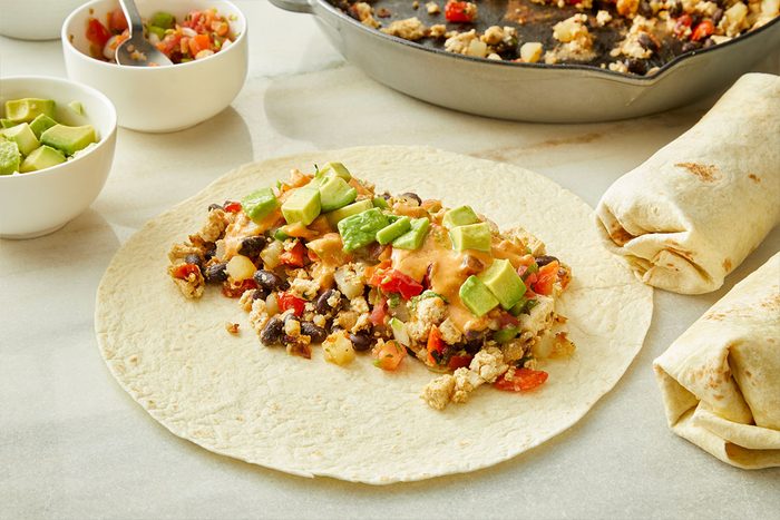 A flour tortilla topped with scrambled eggs, black beans, diced vegetables, and avocado sits on a counter. Nearby are rolled burritos, a skillet with filling, and bowls of avocado and filling.