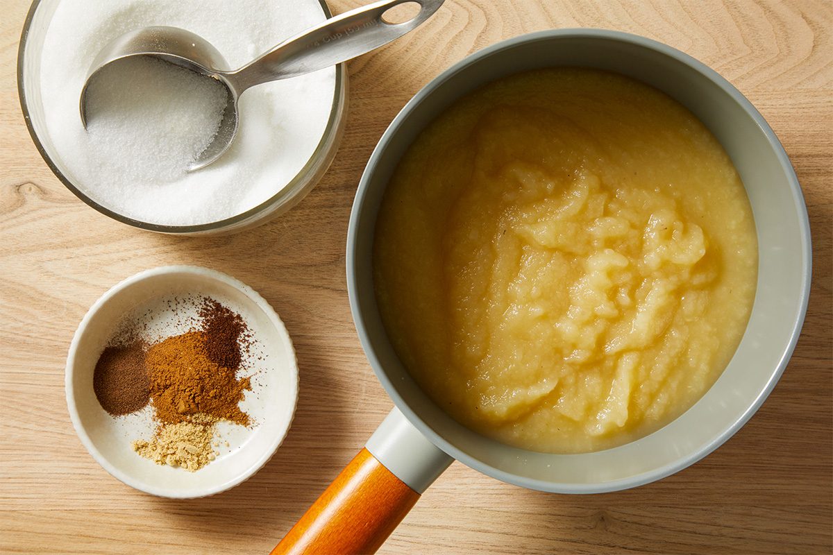 A saucepan with smooth applesauce sits on a wooden surface next to a bowl of granulated sugar with a spoon and a small dish containing four piles of different ground spices.