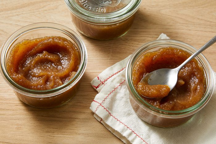 Three glass jars of thick, brown apple butter sit on a wooden surface. One jar has a spoon in it, resting on a folded white cloth with red stitching.