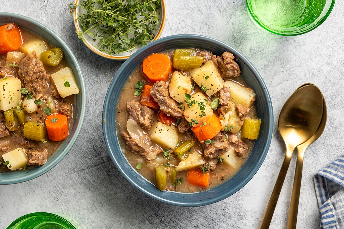 A bowl of beef stew with chunks of beef, potatoes, carrots, celery, and herbs, served on a gray surface with a gold spoon and fork nearby and a small bowl of fresh herbs on the side.