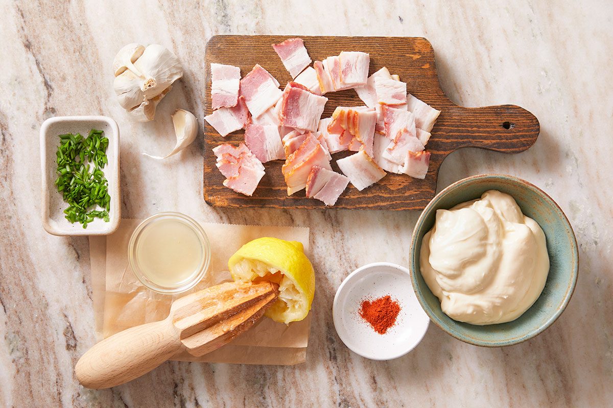 Vertical shot of the raw ingredients for Bacon Aioli, bacon, garlic, lemon, mayonnaise, spices, and chives styled on a wooden board and ceramic bowls