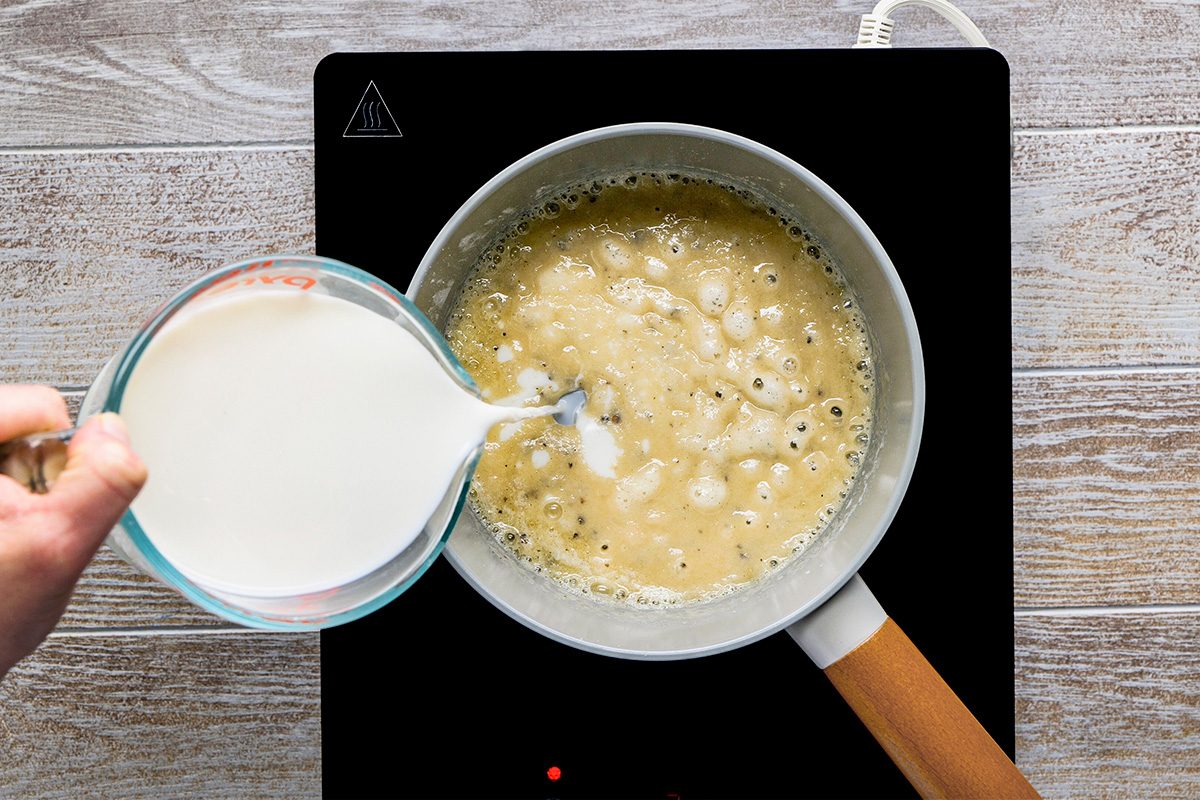 A hand pours milk from a measuring cup into a saucepan of bubbling roux on an electric stovetop, preparing a sauce. The scene is set on a light wooden surface.