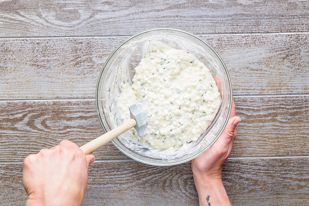 A person holding a glass bowl filled with a creamy, chunky mixture and stirring it with a white spatula, on a wooden surface.