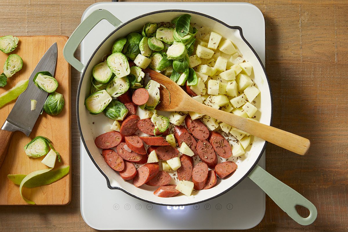 A white pan on a stovetop contains sliced sausage, halved Brussels sprouts, and diced potatoes being cooked, with a wooden spoon inside. A cutting board with a knife and more vegetables is beside the pan.