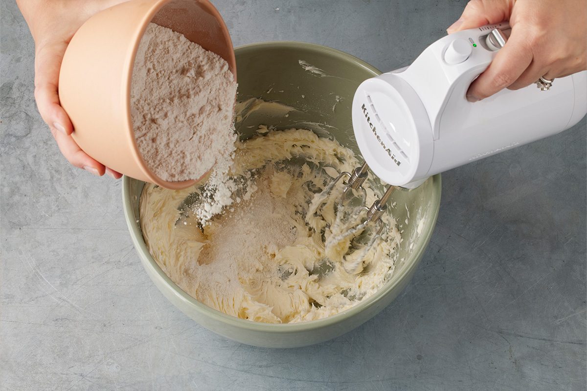 Overhead shot of a person pouring flour from a bowl into a green mixing bowl containing creamed ingredients, while using a white electric hand mixer on a gray countertop;