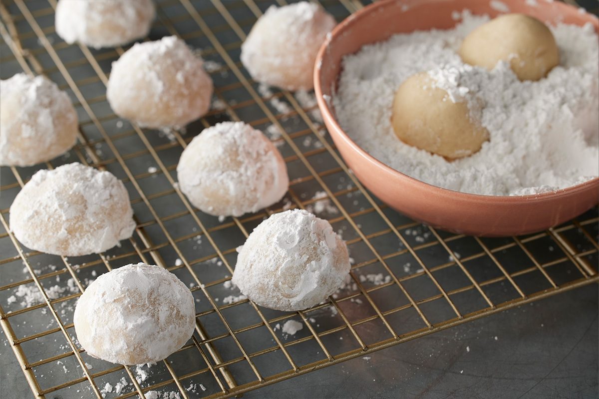 3/4 angle view shot of powdered sugar-coated cookies resting on a cooling rack, with two uncoated dough balls in a bowl of powdered sugar nearby;