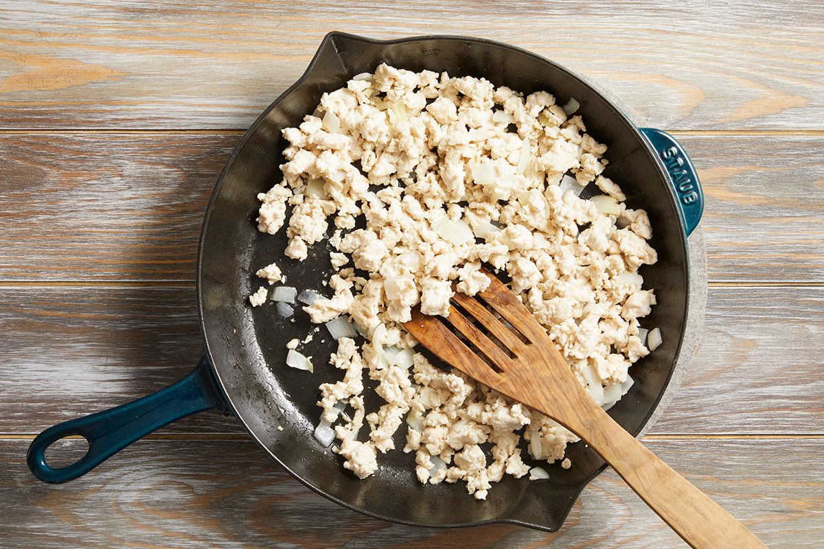 A skillet on a wooden surface contains crumbled tofu being cooked with chopped onions. A wooden spatula rests in the pan, stirring the mixture.