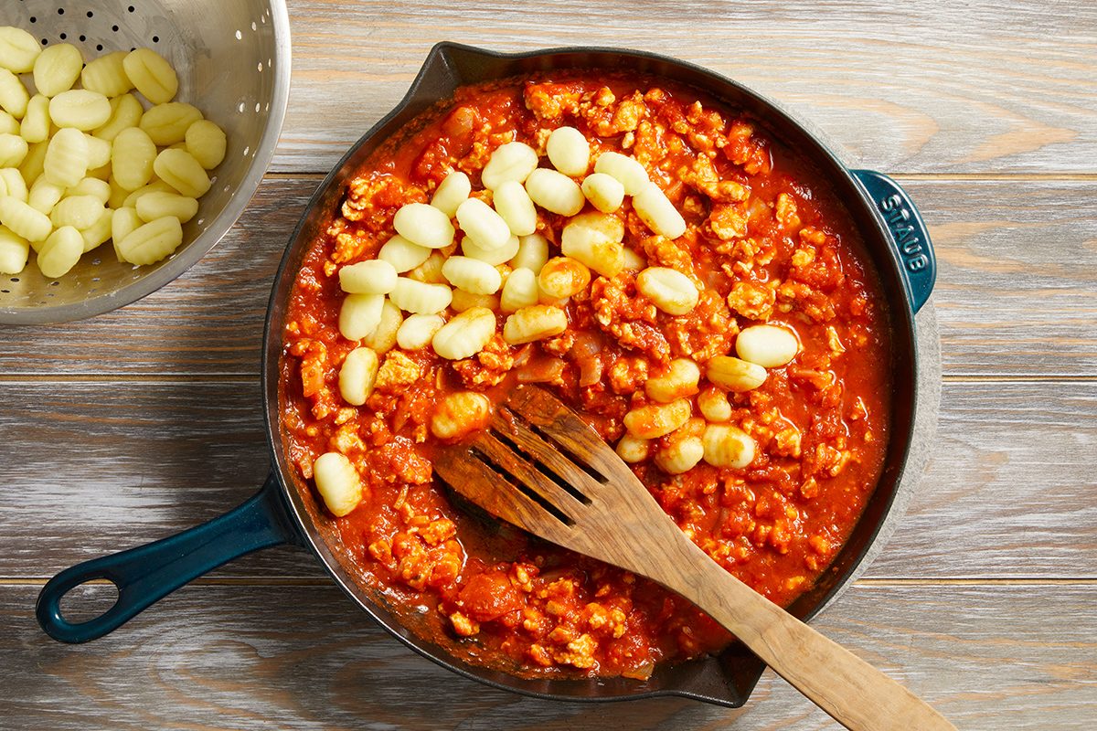 A skillet filled with tomato and meat sauce is topped with gnocchi, and a wooden spoon rests inside. To the side, a metal colander holds more uncooked gnocchi on a wooden surface.