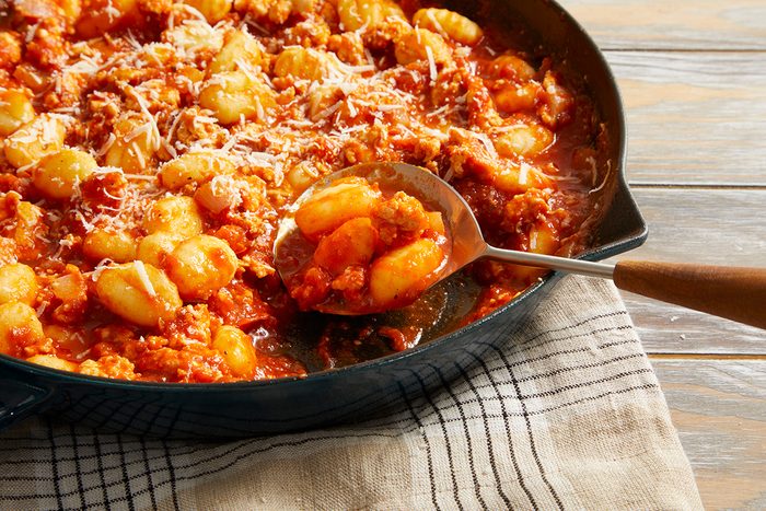 A skillet filled with gnocchi in a rich tomato sauce with crumbled sausage, topped with grated Parmesan cheese. A spoon is scooping some of the dish, resting on a striped cloth.