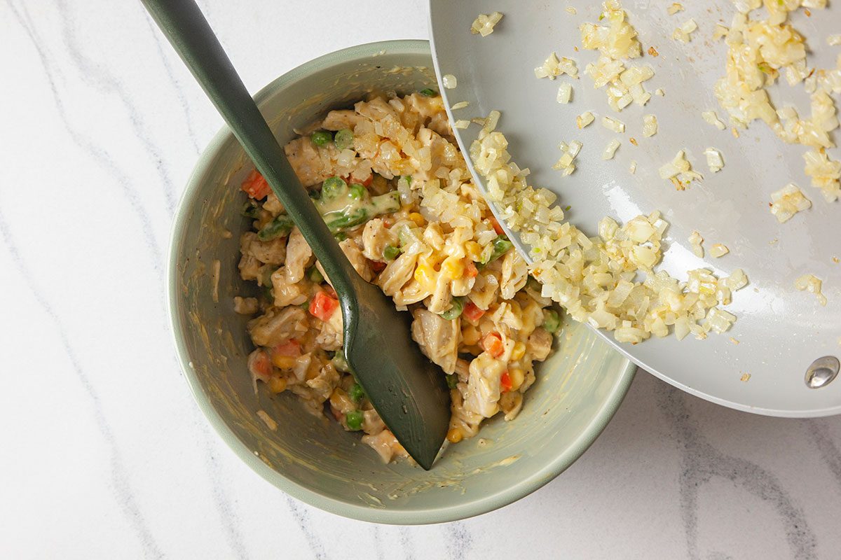 Overhead shot of a bowl of mixed vegetables and chicken being stirred with a dark spoon while sautéed onions are poured in from a pan on a white marble surface;