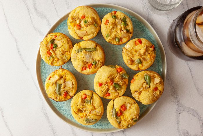 Overhead shot of ten Chicken Potpie Muffins with green beans, red peppers, and other vegetables arranged in a circle on a light blue plate, with a glass of iced drink and a pitcher nearby;