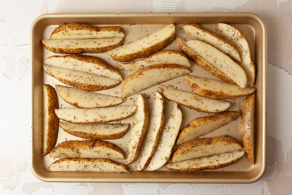 Overhead shot of a baking sheet lined with evenly spaced, seasoned potato wedges with skins on, sprinkled with pepper and herbs, ready to be baked