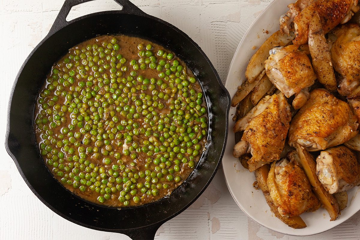 Overhead shot of a skillet of green peas in sauce alongside a platter of seasoned, cooked chicken pieces on a white surface