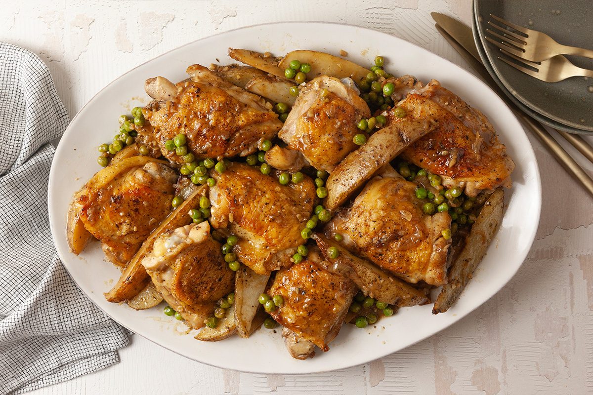 Overhead shot of Chicken Vesuvio on a white platter, featuring roasted chicken thighs, potato wedges, and green peas, set on a light table with gold cutlery