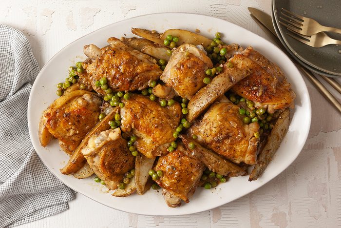Overhead shot of Chicken Vesuvio on a white platter, featuring roasted chicken thighs, potato wedges, and green peas, set on a light table with gold cutlery