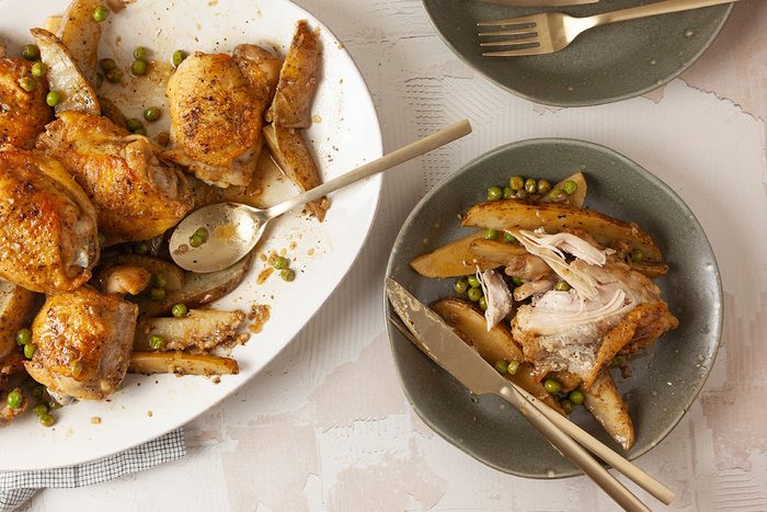 Overhead shot of Chicken Vesuvio served on a large platter and a smaller plate with potato wedges, green peas, utensils, and some shredded chicken on the side