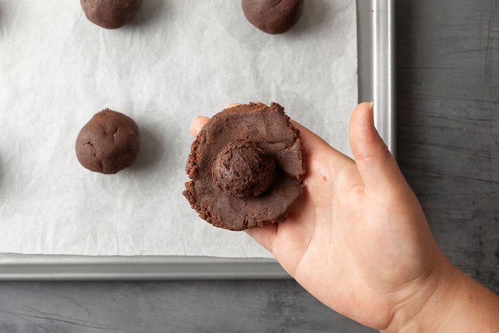 Overhead shot of a hand holds a partially flattened chocolate cookie dough ball above a baking tray lined with parchment paper More round dough balls sit around it The scene shows the cookies being made