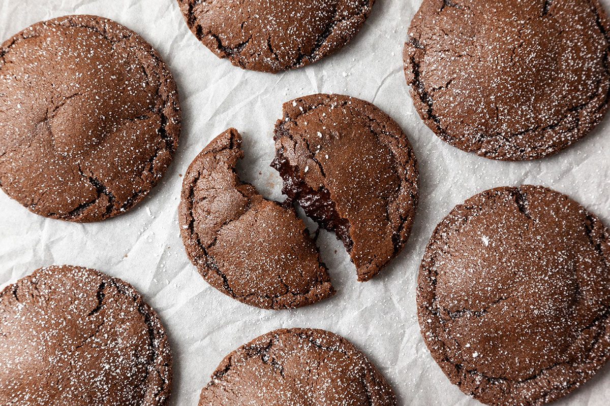 Close-up shot of Chocolate Lava Cookies are laid out on parchment paper and dusted with powdered sugar one cookie in the center is split open showing a gooey inside