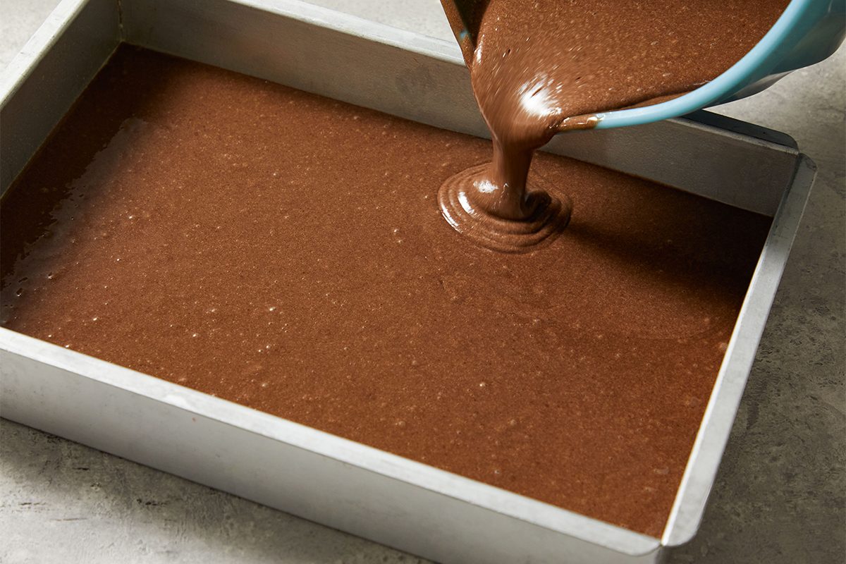 A person pours chocolate cake batter from a blue bowl into a rectangular metal baking pan on a gray countertop.