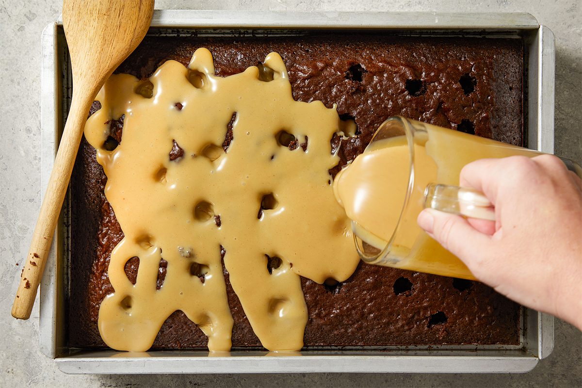 A hand pours caramel sauce from a glass measuring cup over a chocolate sheet cake with holes, using a wooden spoon nearby for spreading. The cake is in a metal pan on a light countertop.