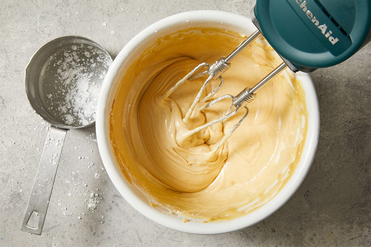 A hand mixer blends creamy batter in a white bowl, with a metal measuring cup containing powdered sugar nearby on a gray countertop.