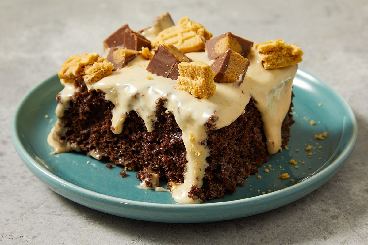 A slice of moist chocolate cake with creamy icing, topped with chunks of chocolate, peanut butter cups, and cookie pieces, served on a turquoise plate.