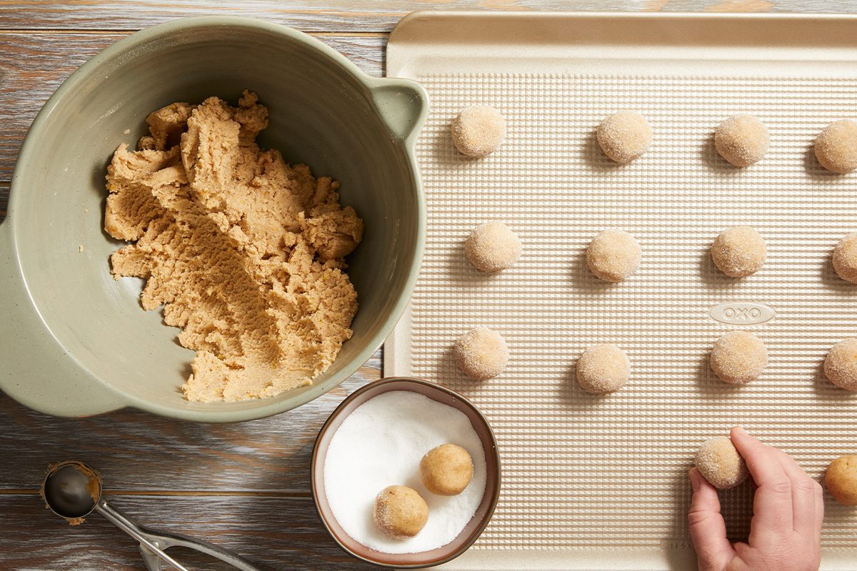 Overhead view of a person shaping cookie dough into balls and placing them on a parchment-lined baking sheet beside a bowl of sugar and cinnamon.