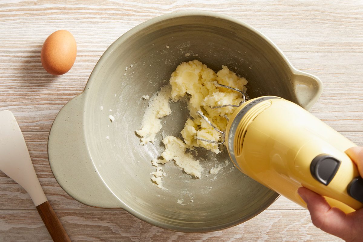 Overhead shot of butter and sugar being creamed with a hand mixer in a mixing bowl with an egg on side