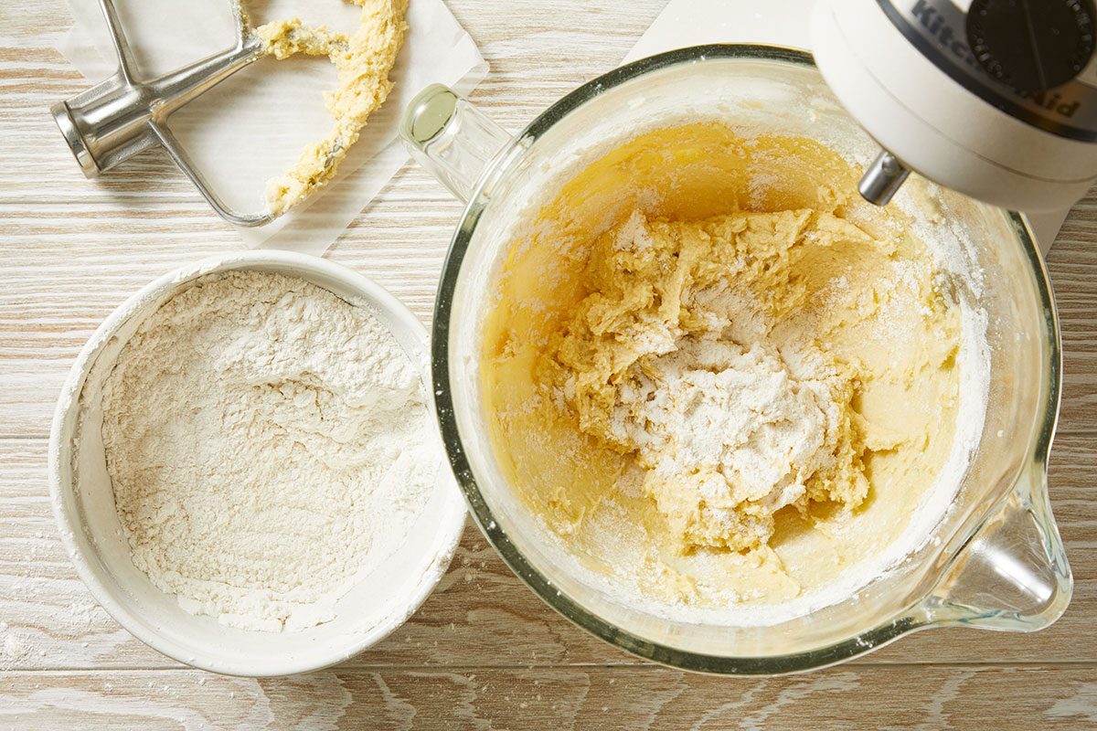 Overhead shot of a stand mixer with creamed dough and a bowl of flour are on a light wooden surface The mixer's paddle attachment lies nearby