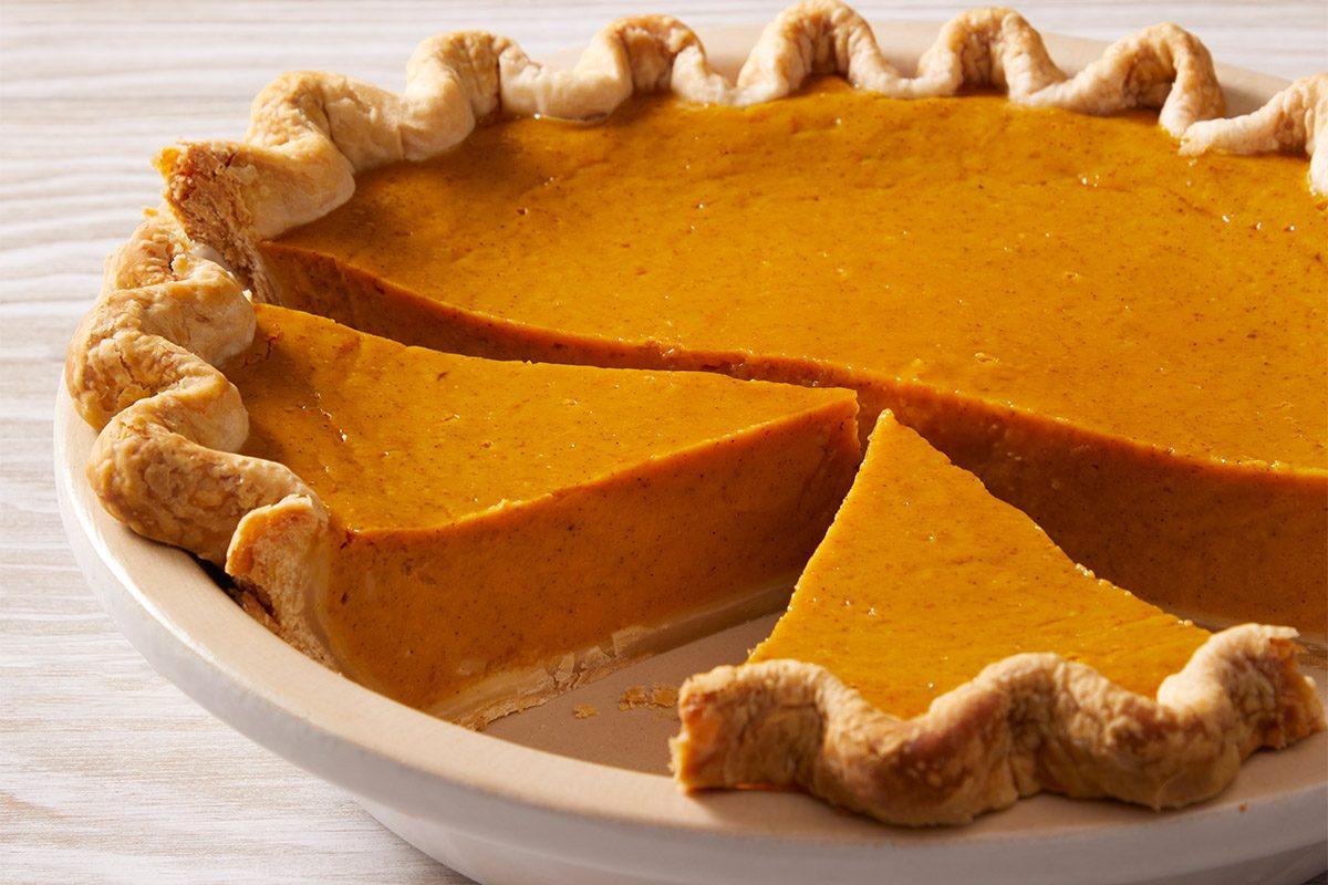 Overhead view of a whole Eggnog Pumpkin Pie with three slices missing; the smooth, spiced filling contrasts with the golden, crimped crust, and a dollop of whipped cream on one slice in the background.
